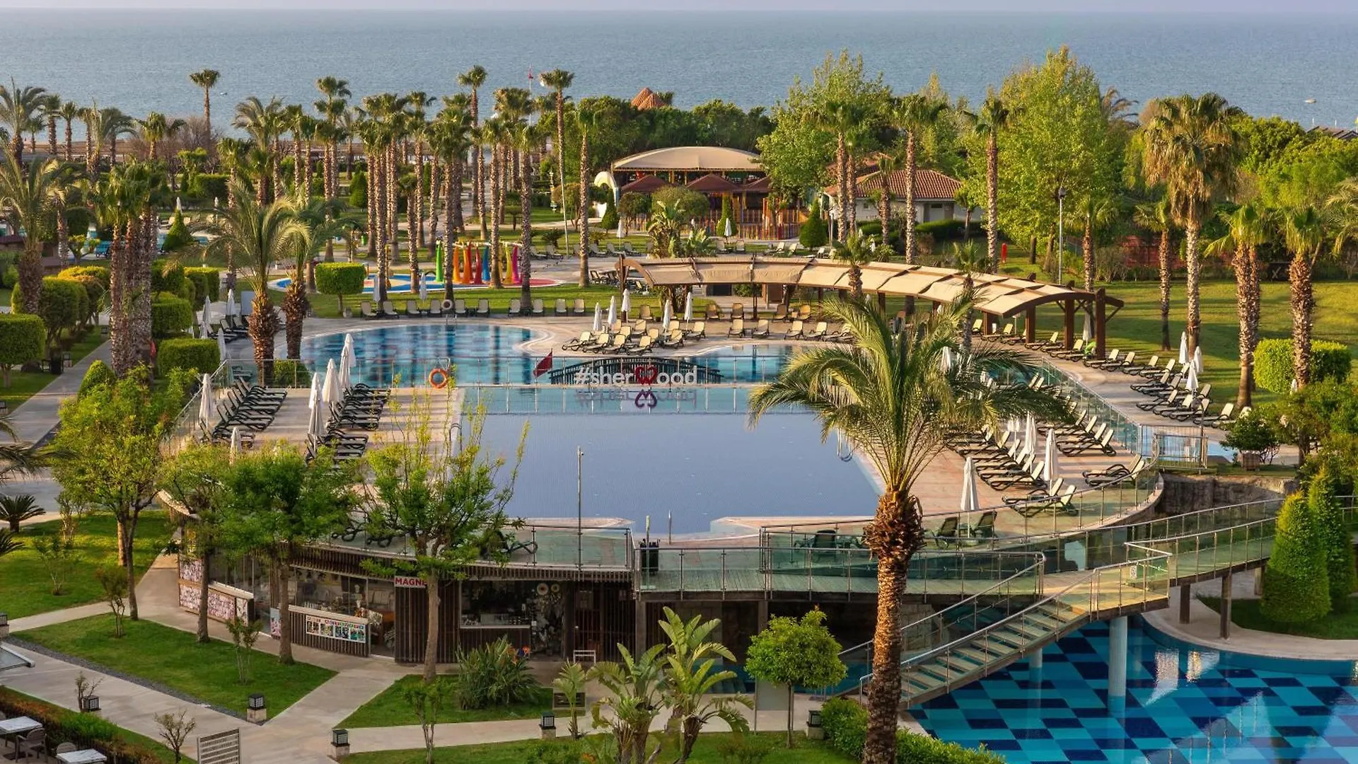 Sherwood Exclusive Lara Antalya Exterior photo The photo shows a beautiful resort area with a large, inviting swimming pool as the central focus. The pool is surrounded by a spacious deck area with numerous lounge chairs, some occupied by people relaxing. 

The resort is situated near a coastline